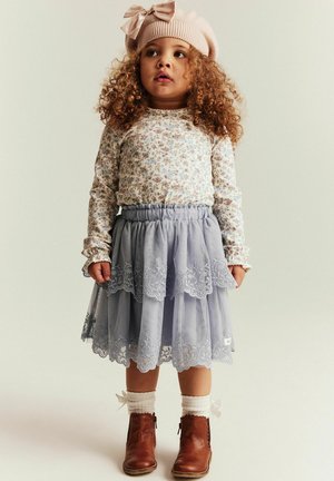 Kleinkind mit lockigem Haar, trägt einen rosa Hut mit Schleife, ein langärmeliges Oberteil mit Blumenmuster, einen grauen Spitzenrock, weiße Socken und braune Stiefel, steht still.