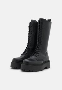 Bottes noires jusqu'aux genoux en cuir lisse, dotées d'une semelle épaisse, d'un laçage à l'avant et de détails de couture le long de la semelle.