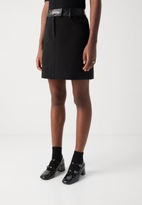 Jupe noire trapèze avec ceinture texturée en similicuir ; associée à des chaussures à talons noires brillantes et des chaussettes noires jusqu'à la cheville. Texture de tissu lisse.