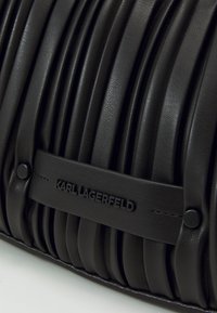 Musta nahkalaukku, jossa on tikattu kuvio ja pystysuuntaiset laskokset. Mukana teksturoitu olkahihna, jossa on logo "KARL LAGERFELD". Sileä viimeistely.