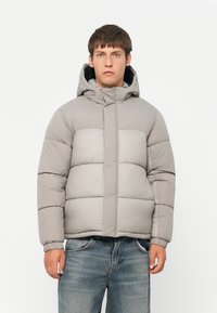 Polstret grå jakke med hette, med et quiltet design, elastiske mansjetter og glidelås foran, båret over slitte jeans.