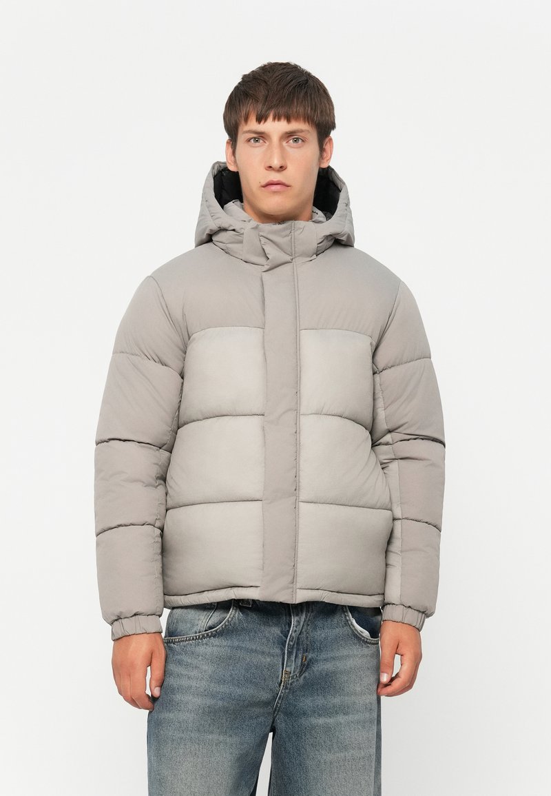Polstret grå jakke med hette, med et quiltet design, elastiske mansjetter og glidelås foran, båret over slitte jeans.