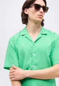 Νεαρός άνδρας με σκούρα γυαλιά ηλίου και φωτεινό πράσινο πουκάμισο με κοντά μανίκια και κουμπιά, που κοιτάζει προς τα πλάγια με σταυρωμένα τα χέρια.