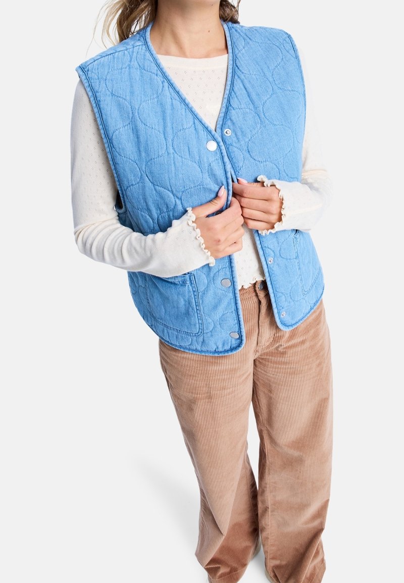 Lichtblauwe gewatteerde bodywarmer met knoopsluiting, een gladde textuur en afgeronde randen, gedragen over een wit shirt met lange mouwen en een broek van ribfluweel.