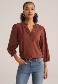 Blouse marron texturée avec un col volanté, boutonni�ère à l'avant et manches bouffantes 3/4, associée à un jean bleu et une ceinture noire.