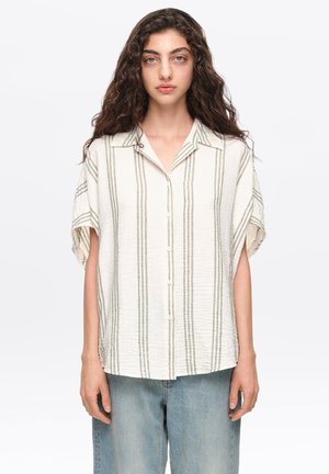 Mujer con cabello largo y rizado que lleva una camisa blanca de manga corta y suelta con rayas verticales grises y vaqueros de tiro alto de color azul claro.