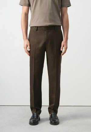 Mężczyzna ubrany w ciemnobrązowe, dopasowane spodnie, beżową koszulę z krótkim rękawem oraz czarne skórzane mokasyny, stojący na tle jasnego, jednolitego tła.