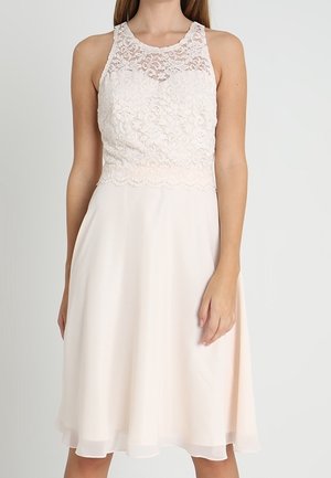 Žena v bezrukávých bílých šatech po kolena s krajkovým živůtkem a vzdušnou sukní, stojící před jednoduchým světlým pozadím.