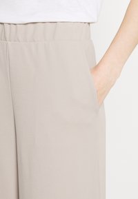 Jasnobeżowe szerokie spodnie z elastycznym pasem i boczną kieszenią, wykonane z gładkiego, miękkiego materiału. Prosty design.