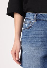 Denim mini suknja s izblijedjelom plavom završnom obradom, koja se ističe prednjim džepom, ispucalim detaljima i lagano frženim rubom. Crni top djelomično vidljiv.