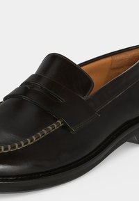 Μαύρο δερμάτινο penny loafer με ορατή ραφή και ταμπά εσωτερική επένδυση, εμφανιζόμενο από κοντινή πλευρική γωνία.