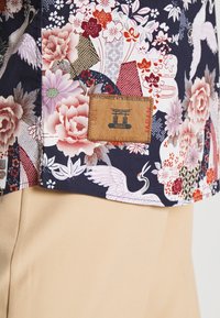 Patroontekstiel met bloemenmotieven, kraanvogels en geometrische vormen in marineblauw, roze en rood. Inclusief een leren label met de tekst "INDIGO".