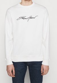 Vit sweatshirt med rund hals, ribbade ärmar och nederkant. Har svart textlogotyp i kursiv stil över bröstet. Slen tygtextur.