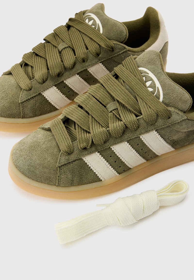 Sneaker in suede verde oliva con tre strisce bianche, lacci testurizzati e suola in gomma. Include un ulteriore laccio di colore crema.