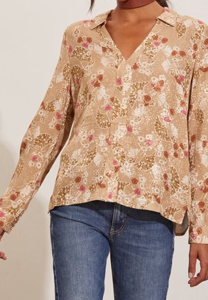 Blouse beige à manches longues avec un motif floral rose, blanc et marron, portée avec un jean en denim bleu et un collier ras du cou sombre.