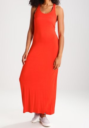 Kobieta ubrana w długą, pomarańczową sukienkę bez rękawów oraz jasnoróżowe wsuwane buty, stojąca na tle jednolitego jasnego tła.