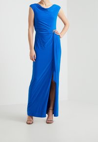 Blue maxi dress made of smooth fabric, featuring a cowl neckline, asymmetrical front drape, side slit, and decorative hardware accent.