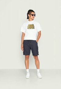 Ung man med solglasögon som bär en vit grafisk t-shirt, svarta cargoshorts, vita strumpor och vita sneakers, stående mot en neutral vägg.