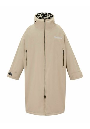 Kobieta w beżowym długim płaszczu z podszewką w zwierzęcy wzór, beżowym dzianinowym swetrze, białych spodniach, czarnych butach i brązowej dzianinowej czapce, pozująca na jasnym tle.