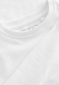Hvid bomulds t-shirt med en glat tekstur, rund ribbet halsudskæring og mærkedetalje synlig indvendigt, lagt fladt mod en anden skjorte.
