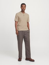 Mand stående iført en beige kortærmet polo, grå pressede bukser og brune lædersko foran en ensfarvet baggrund.
