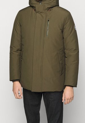 Mann trägt eine olivgrüne gesteppte Jacke mit Kapuze, Reißverschluss-Brusttasche und Seitentaschen, kombiniert mit dunkler Hose vor einem einfarbigen Hintergrund.