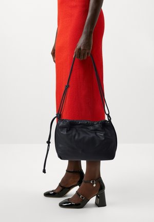 Personne en robe rouge tenant un sac à main en cuir noir, portant des escarpins Mary Jane en cuir verni noir avec brides à la cheville.