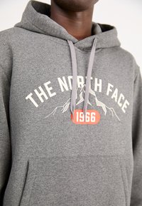 Szara bluza z kapturem wykonana z miękkiego materiału, z przednią kieszenią, sznurkami oraz graficznym nadrukiem z górami i napisem: "THE NORTH FACE 1966."