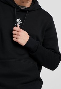 Felpa nera con cappuccio, tasca a marsupio, polsini a coste e coulisse regolabili. Presenta un piccolo logo bianco sul petto. Tessuto dalla texture morbida.