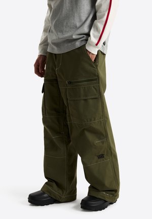 Man met olijfgroene wijde cargobroeken met zijzakken, zwarte schoenen en een grijs shirt met lange mouwen met een rode streep op de mouw.