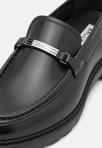 Melni ādas loafers ir ar gludu virsmu, bezvīļu dizainu un ar sudraba toņa metāla akcentu, uz kura ir markas nosaukums "Bally".
