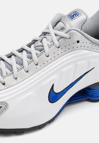 Sivi i bijeli tenisice s teksturiranim gornjim dijelom, plavim naglascima, iznimnim Nike swoosh logom i vidljivim Shox amortizerom u peti.