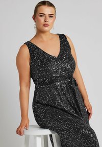 Vestido de noche negro con lentejuelas, con escote en V, sin mangas, cintura ceñida y falda fluidas. La tela tiene un aspecto brillante y texturizado.