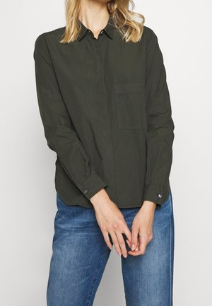 Femme portant une chemise boutonnée manches longues vert foncé avec une poche poitrine et un jean bleu, debout devant un fond uni.