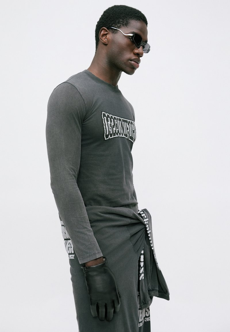 Mand iført en mørkegrå langærmet skjorte og bukser med hvid tekst, sort læderhandske på venstre hånd og sorte solbriller mod en hvid baggrund.