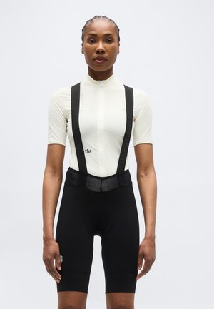 Frau trägt ein weißes Radsport-Trikot mit kurzen Ärmeln und schwarze Trägerhose, steht mit dem Gesicht zur Kamera vor einem schlichten Hintergrund.