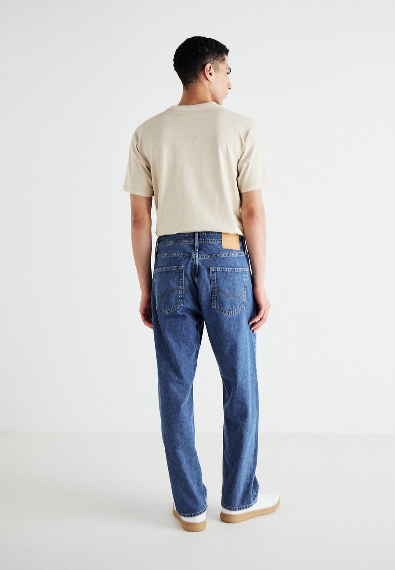 T-shirt beige chiaro con maniche corte, abbinato a jeans in denim blu con due tasche posteriori e una toppa in pelle beige in vita. Sneakers bianche.