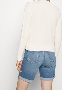 Cremfarbener, gestrickter Pullover mit strukturierten Ärmeln und geripptem Saum, kombiniert mit hellblauen Jeansshorts mit umgeschlagenen Säumen und Gesäßtaschen.