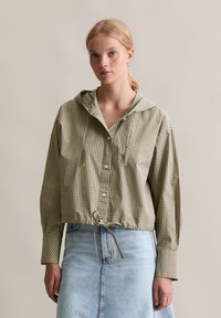 Gekürztes Hoodie-Shirt in olivgrün mit einem weißen Karomuster, mit Knopfverschluss und Kordelzug an der Taille, kombiniert mit einem Jeansrock.