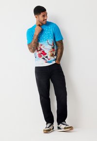 Modré tričko s veselým motivem Santy a soba, obklopené sněhovými vločkami; ve spojení s černými džínami a vícero barevnými teniskami.