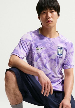 Mladý muž vyladěný v fialovém fotbalovém dresu Jižní Koreje s květinovým vzorem, černých šortkách a šedých ponožkách pózuje s jedním kolenem zvednutým na bílém pozadí.