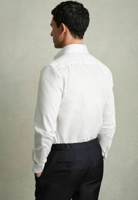 Weißes Hemd mit langen Ärmeln, sichtbarem Kragen und Knopfmanschetten, kombiniert mit dunklen, maßgeschneiderten Hosen mit einer Gesäßtasche.