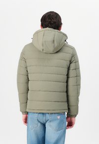 Polstret grønn jakke med hette, med horisontalt quiltet design, elastiske mansjetter og en avslappet passform, kombinert med blå jeans.