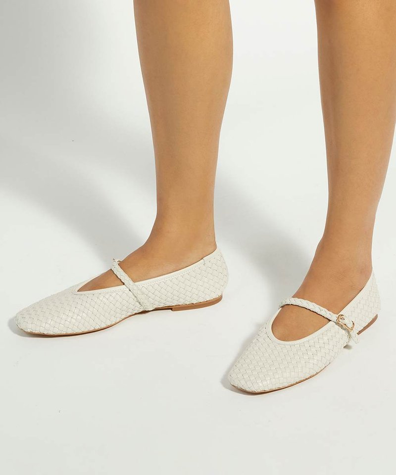 Pieds portant des ballerines Mary Jane blanches tissées avec une fine bride et une petite boucle dorée sur un fond clair.