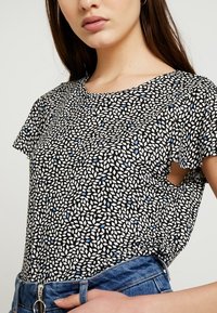 Černobílý vzorovaný top s krátkými rukávy, zdobený malými modrými akcenty, kulatým výstřihem a volným střihem. Měkká textilie.