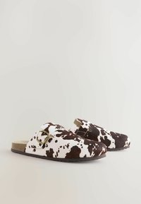 Zuecos de color marrón y blanco con estampado de vaca, con una correa de hebilla, textura similar a fieltro y suela plana de goma. Diseño de puntera redondeada.