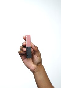 Produkt do ust Revlon Colorstay Suede Ink w dwukolorowym opakowaniu: różowa góra i czarna podstawa, trzymany w dłoni na tle białego, gładkiego tła.