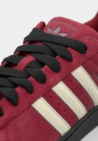 Red suede sneaker with black laces, featuring three white stripes, perforations on the sides, and a gray logo on the tongue.