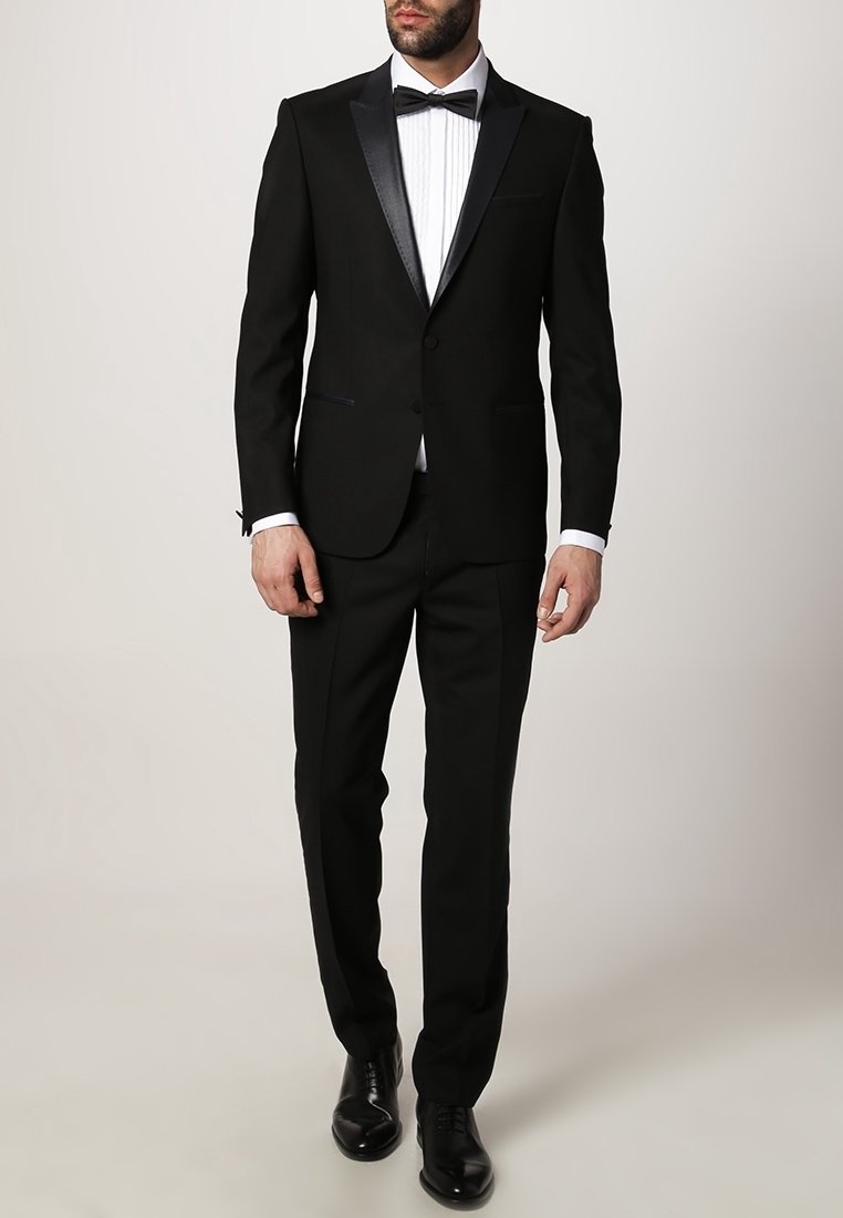 Homme portant un smoking noir avec des revers en satin, une chemise blanche, un nœud papillon noir, un pantalon noir et des chaussures noires, debout devant un fond uni.