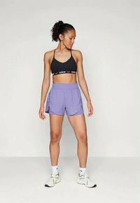 Musta spordibrassi valge logoga, mille juurde on sobitatud heledat lilla sorti lühikesed püksid, mis sisaldavad vöökohta ja küljetaskut. Mudel kannab valgeid spordijalatsisid.